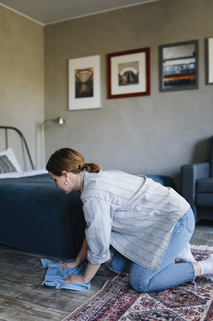 A woman with brown hair tied back, dressed in a light striped shirt and jeans, is kneeling on a patterned rug while cleaning a wooden floor with a blue cloth. She is positioned near a bed with a dark blue bedspread and white pillows, in a contemporary living room with beige walls adorned with three framed pictures. Natural light illuminates the space, highlighting the clean, polished appearance of the wooden surface as part of a domestic surface cleaning and maintenance routine, with the setting emphasizing hygiene and tidy surroundings, typical of professional carpet and floor cleaning services provided by Catford Carpet Cleaners in Catford.