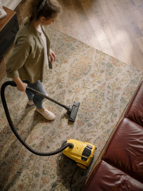 A woman with brown hair tied back, dressed in a light striped shirt and jeans, is kneeling on a patterned rug while cleaning a wooden floor with a blue cloth. She is positioned near a bed with a dark blue bedspread and white pillows, in a contemporary living room with beige walls adorned with three framed pictures. Natural light illuminates the space, highlighting the clean, polished appearance of the wooden surface as part of a domestic surface cleaning and maintenance routine, with the setting emphasizing hygiene and tidy surroundings, typical of professional carpet and floor cleaning services provided by Catford Carpet Cleaners in Catford.