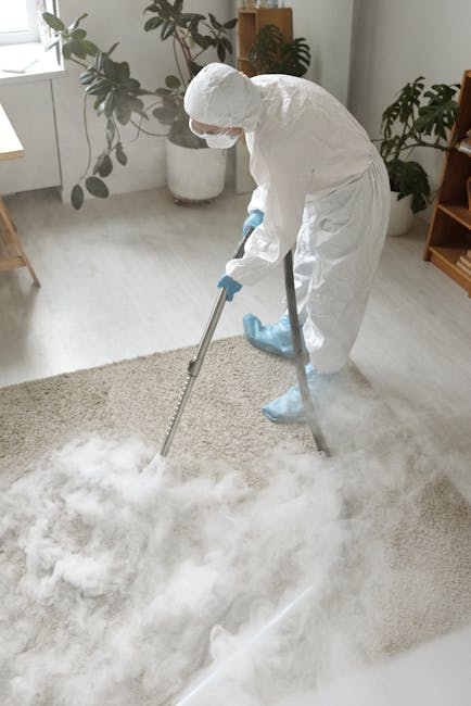 A woman with brown hair tied back, wearing a light blue and white striped shirt and blue jeans, is kneeling on a patterned area rug and using a cloth to clean the wooden flooring in a living room. The