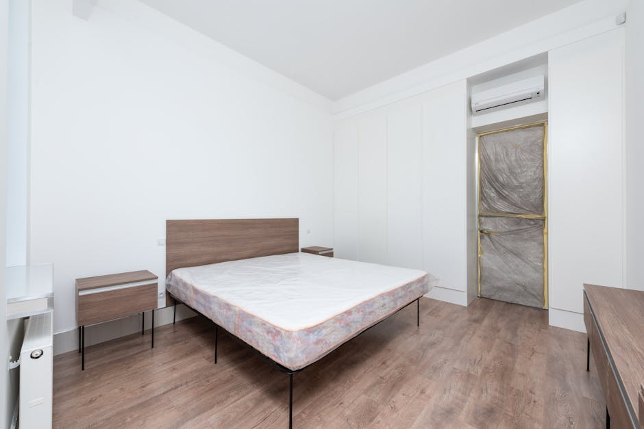A woman with blonde hair wearing a light-colored shirt is leaning over a bed, adjusting a beige bedspread or mattress protector in a well-lit bedroom setting. The room features a white wall, a small w